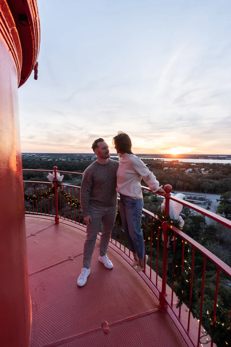 Eric and Anastasia share an intimate moment on a red observation tower at sunset, gazing at each other against a scenic landscape backdrop.