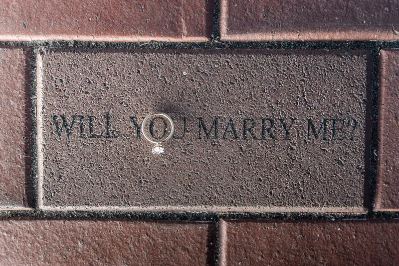 A close-up detail shot of an engagement ring placed on engraved brick pavement that reads 'WILL YOU MARRY ME'.