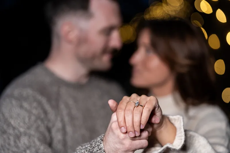 An intimate engagement photo showcasing the couple's clasped hands with a sparkling diamond ring, while they gaze lovingly at each other in soft focus.