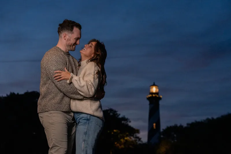 Eric and Anastasia share an intimate embrace during golden hour with a illuminated lighthouse glowing in the background during their engagement session.
