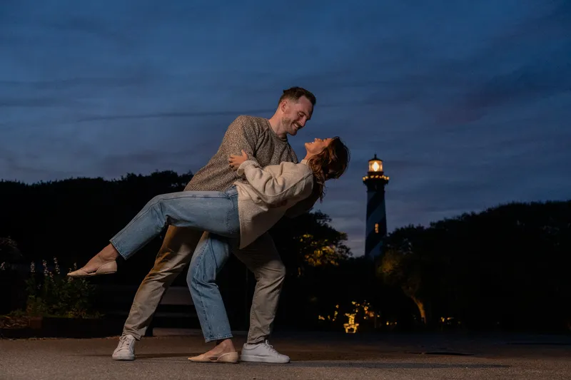 Eric dips Anastasia in a romantic pose during their engagement session at dusk with a lighthouse glowing in the background.