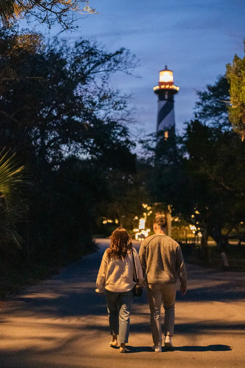 Eric and Anastasia walk hand in hand down a tree-lined path at dusk, with a beautifully lit lighthouse glowing in the background.