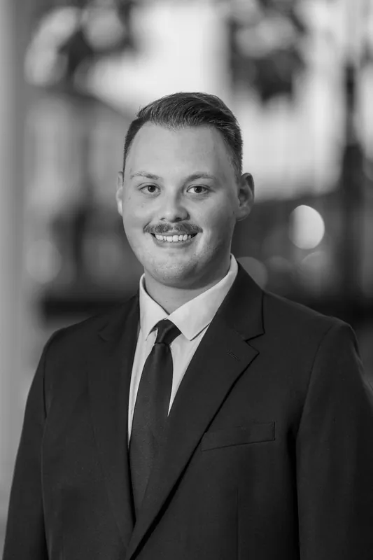 Black and white portrait of Eric in a dark suit and tie, smiling with historic architecture softly blurred behind him.