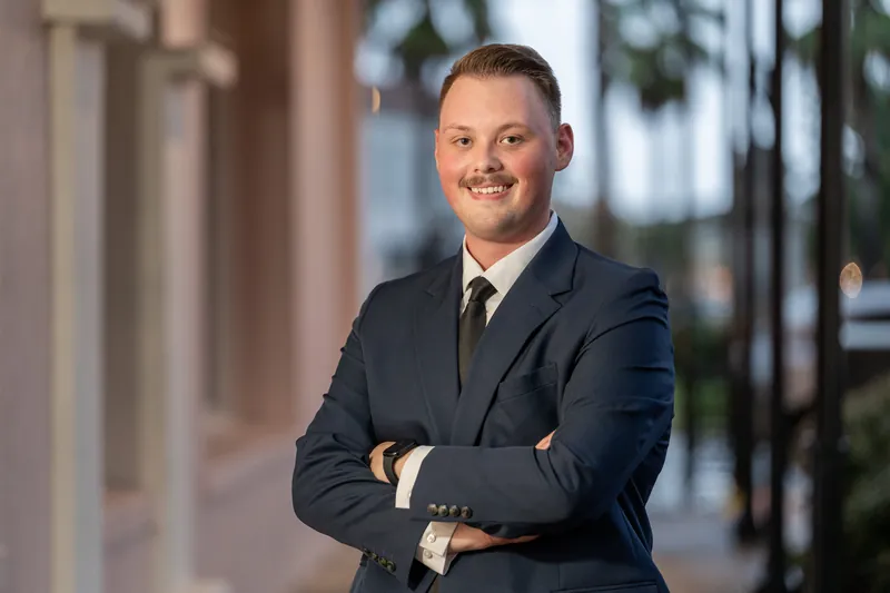 Eric stands with arms crossed, wearing a navy blazer and tie, with colonial columns and greenery framing the scene.
