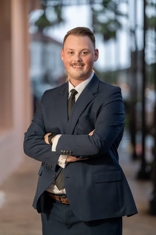 Eric poses with crossed arms in a navy suit on a Downtown St Augustine sidewalk with soft warm lighting.