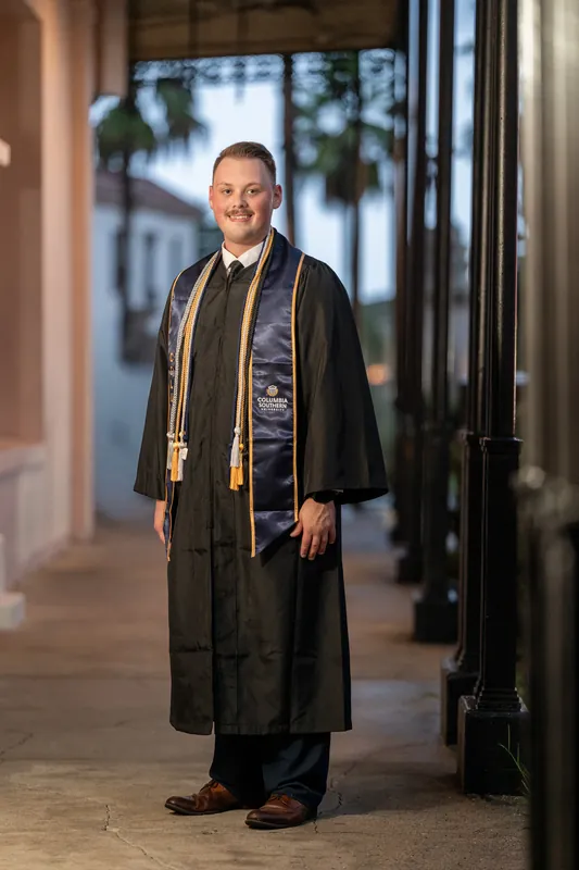 Eric wears a black graduation gown and stole over his suit, standing in the colonnade of a historic building.