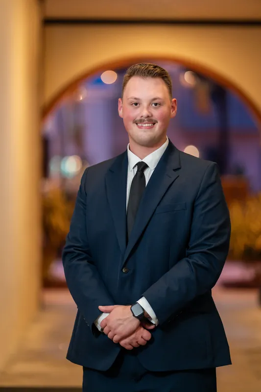 A person in a dark suit and tie smiles with hands clasped in front of an arched doorway at Downtown St Augustine.