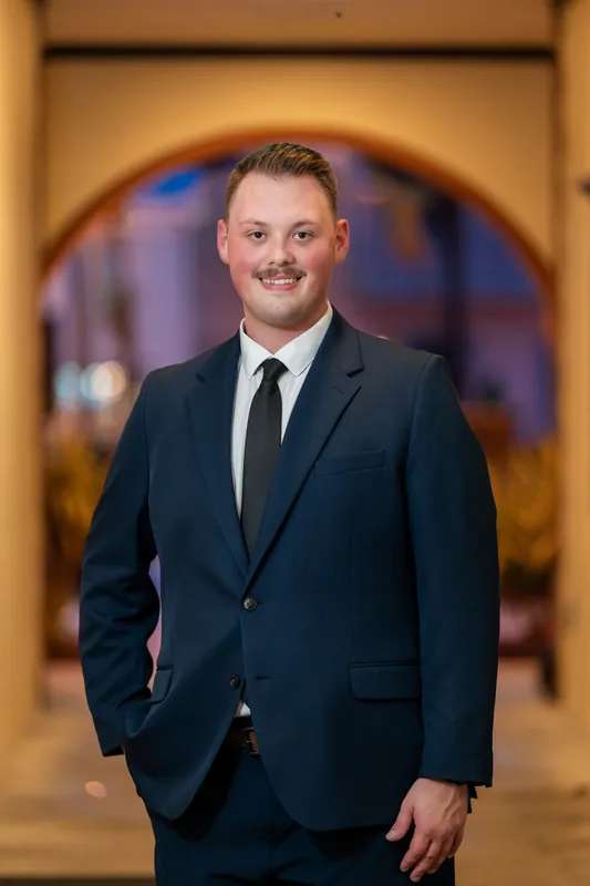 A person in a navy blazer and tie stands with hand in pocket beneath a warm-lit arch at Downtown St Augustine.