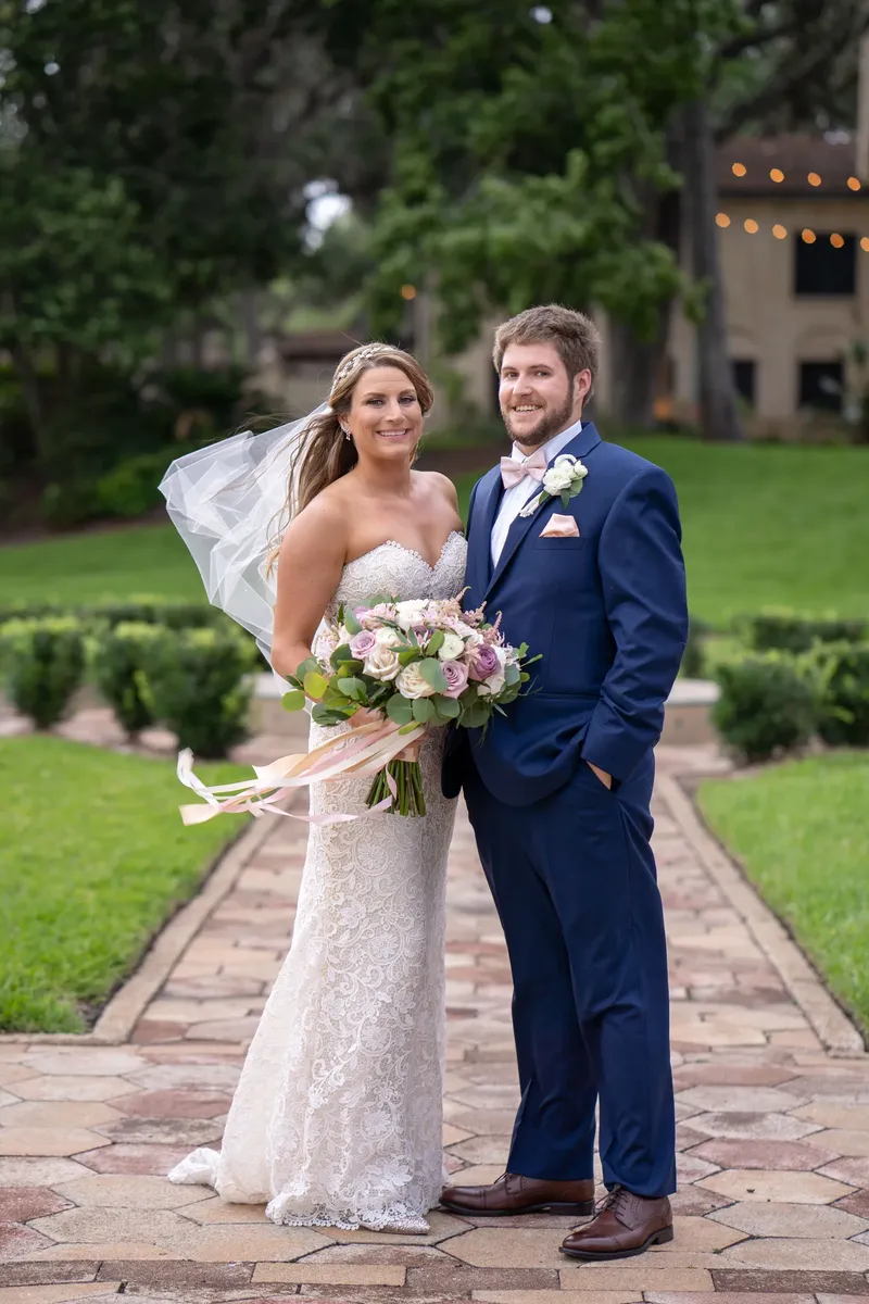 Erin and Sam stand on a brick pathway, Erin in a lace gown with veil holding a white and blush bouquet, Sam in a navy suit.
