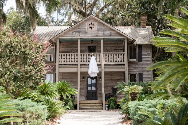 A person in a white wedding gown stands on the front porch of a historic wooden house surrounded by lush greenery at La Venture Grove.