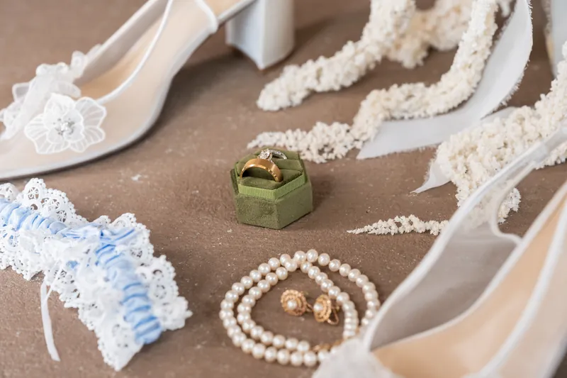 White heels, a blue garter, pearl strands, and two gold rings arranged on a neutral surface with white lace details.