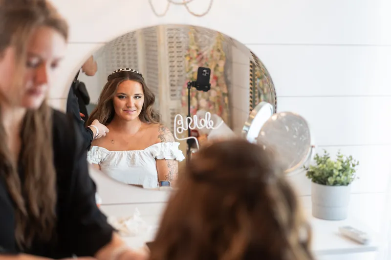 A person in an off-shoulder white wedding dress smiles in a round mirror while friends assist with final preparations.
