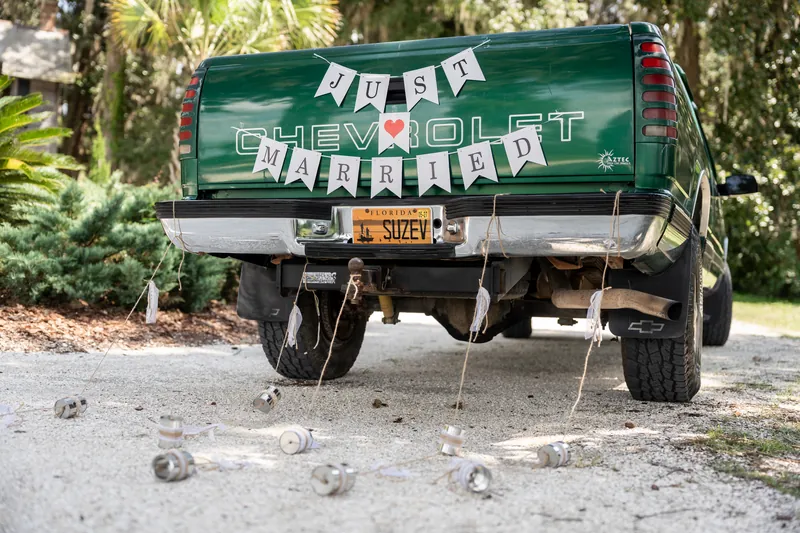 A vintage green Chevrolet truck decorated with "JUST MARRIED" and "CHEVROLET" banners parked on a tree-lined driveway.