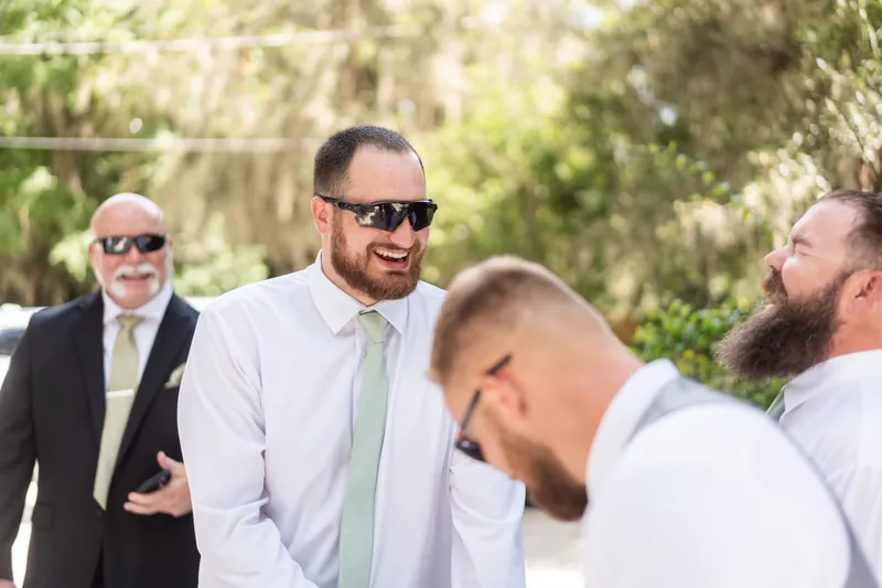 A group of men in light-colored suits and ties laugh together outdoors surrounded by Spanish moss-draped trees.