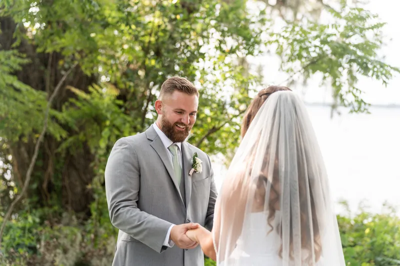 Under dappled tree shade at La Venture Grove, a person in a gray suit holds hands with their partner wearing a white wedding gown and veil.
