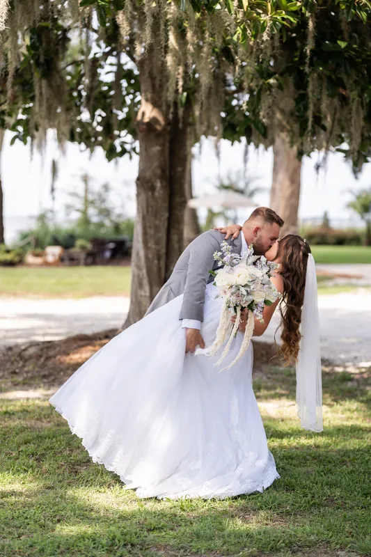 Evelynn and Kevin share a kiss beneath a sprawling oak tree at La Venture Grove, with white flowers visible in the foreground.
