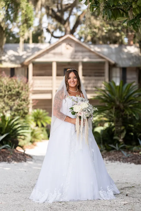 Evelynn stands in a white lace gown and veil, holding a white bouquet in front of a cottage at La Venture Grove, St. Augustine.