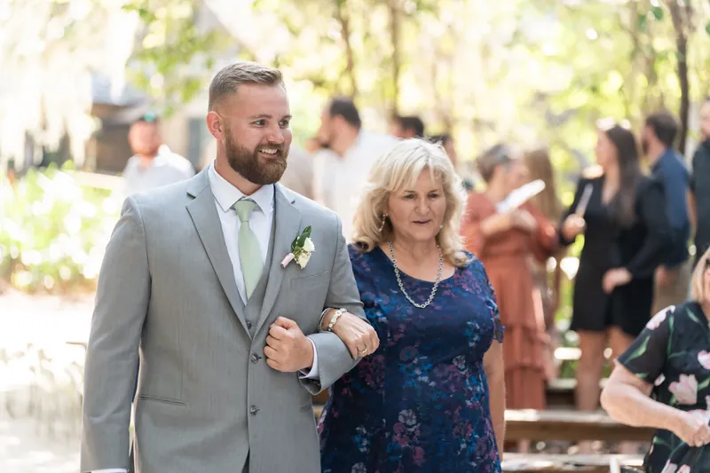A person in a gray suit walks arm-in-arm with a woman in a navy floral dress through the tree-lined grounds.