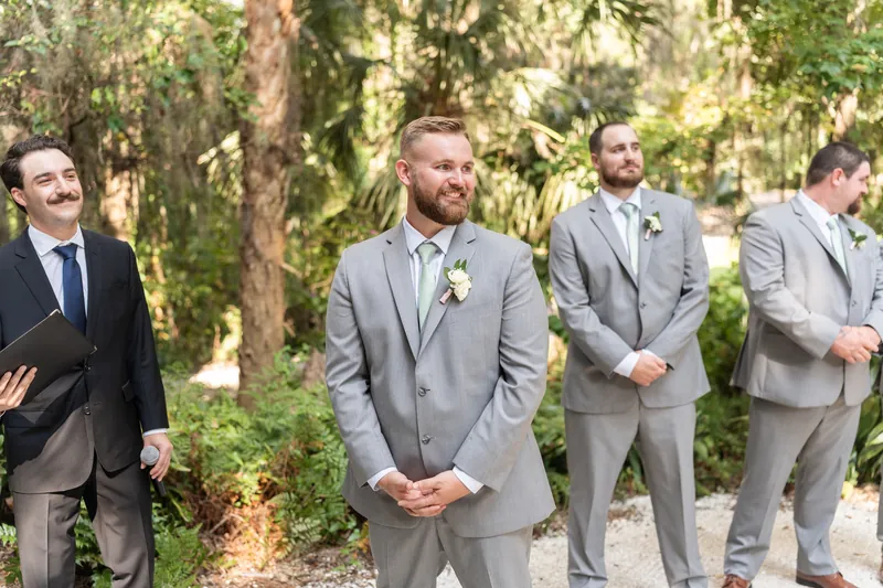 Kevin stands in a gray suit with three groomsmen in matching attire along a shaded pathway lined with Spanish moss.