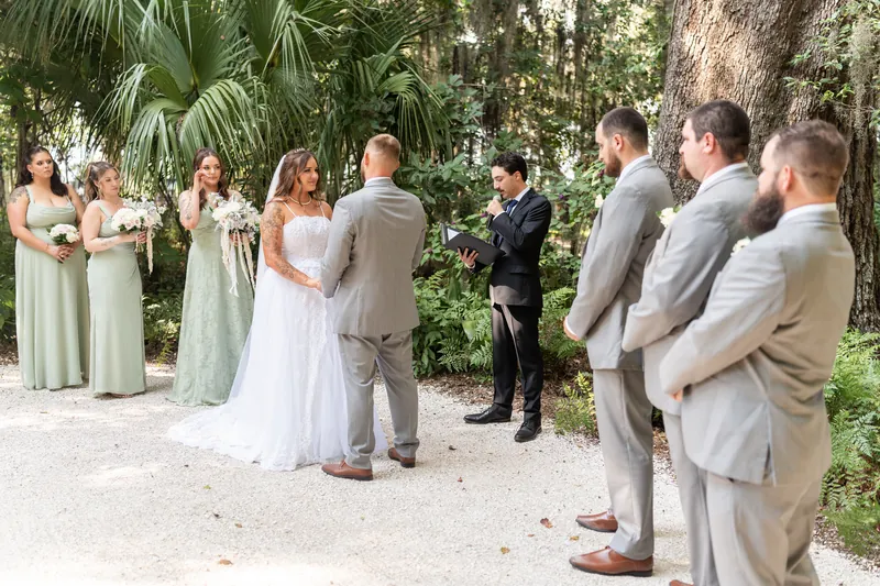 The couple faces each other during their ceremony beneath Spanish moss trees, surrounded by bridesmaids and groomsmen in soft pastels and gray.