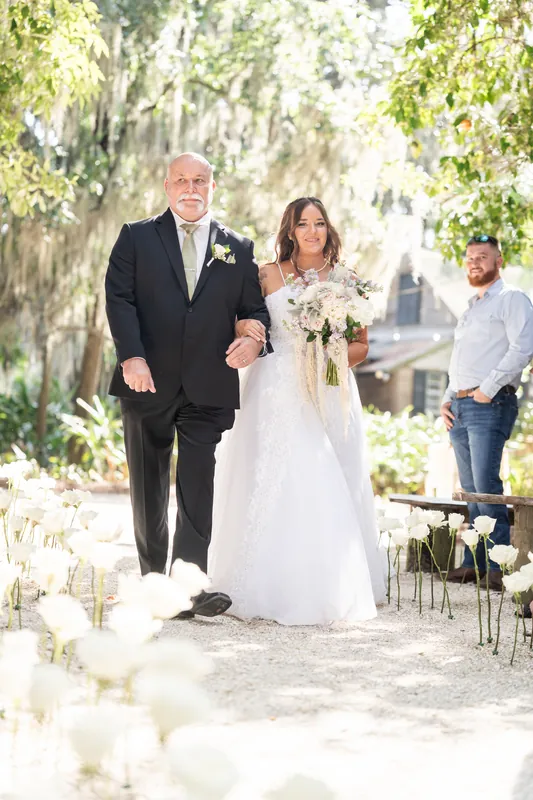 A person in a dark suit walks Evelynn down the aisle as she carries a cream bouquet, white lanterns flanking the path.