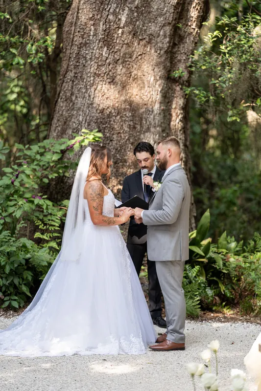 Kevin places a ring on Evelynn's finger while an officiant looks on during their ceremony under the sheltering oak.