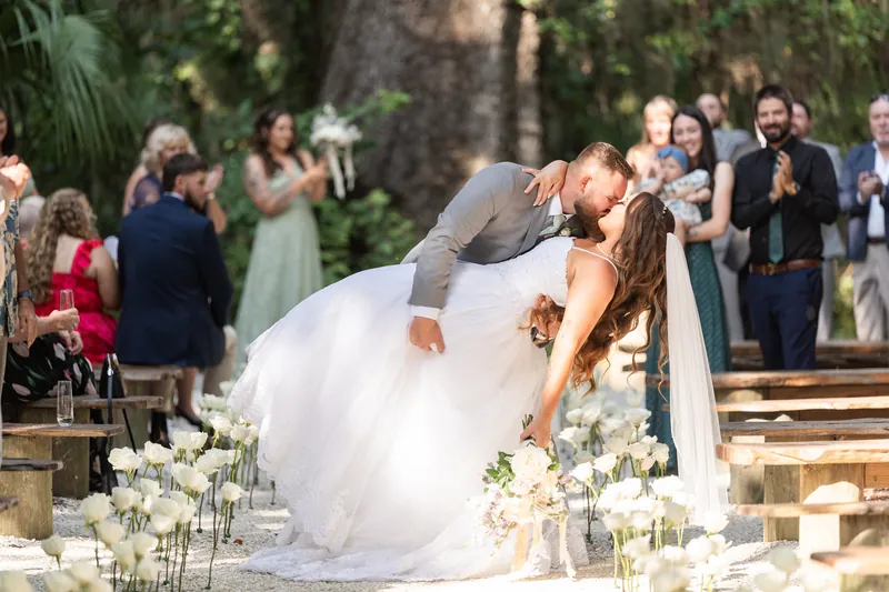 Evelynn and Kevin kiss as their wedding party and guests applaud around white floral arrangements at La Venture Grove.
