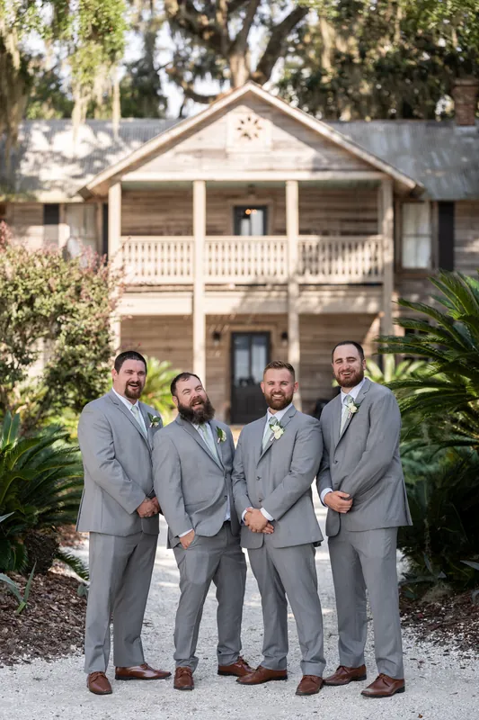 Four groomsmen in gray suits stand shoulder-to-shoulder in front of a historic two-story plantation house.