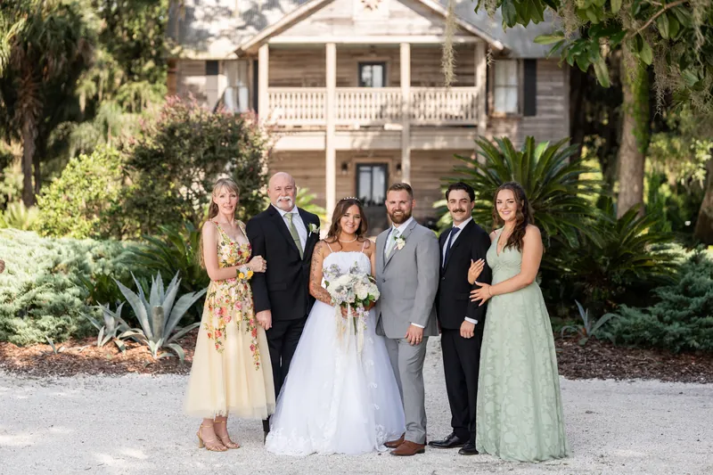 Evelynn in a white gown and Kevin in a gray suit pose with four family members in front of the La Venture Grove mansion.