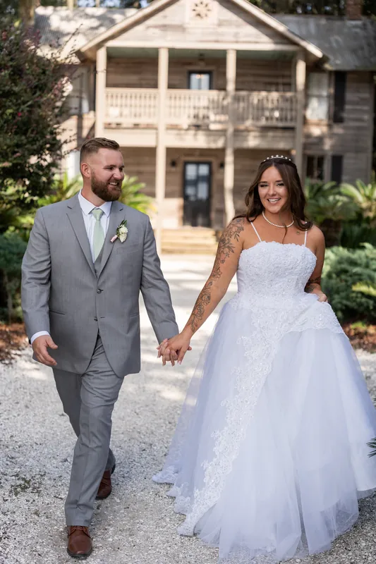 Evelynn and Kevin hold hands while walking on a gravel path in front of a cream-colored mansion at La Venture Grove.