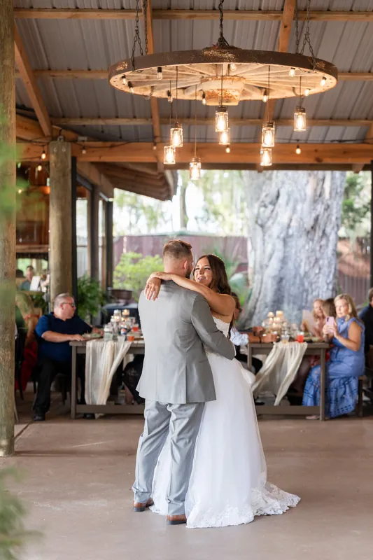 Two people in wedding attire dance together under a circular chandelier in an open-air pavilion surrounded by guests.