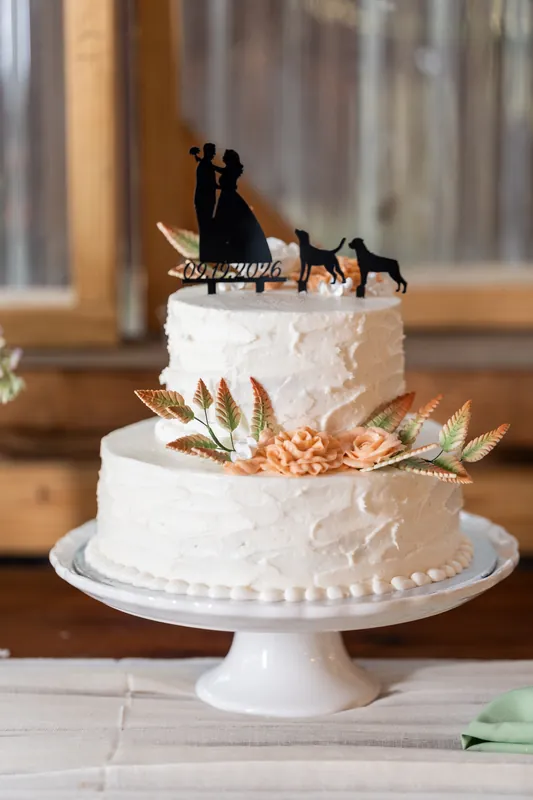 Two-tier white wedding cake topped with a couple figurine and gold wheat accents sits on a pedestal.