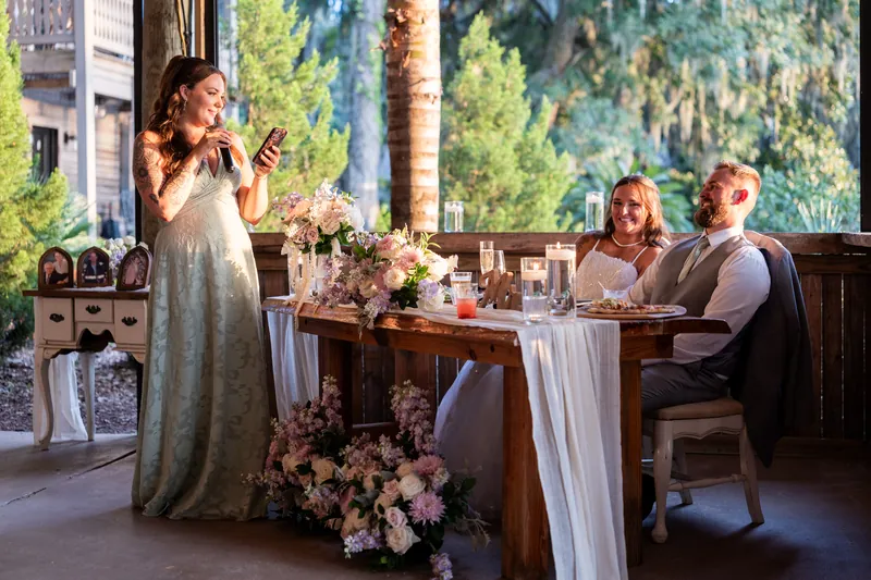 A woman in a sage gown gives a toast while two seated guests listen at a long table decorated with pink florals.