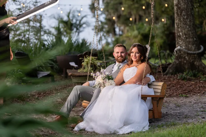Two people in wedding attire sit on a swing strung between trees, framed by greenery and warm evening light.