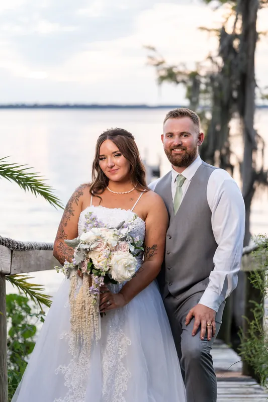 Evelynn and Kevin stand by the water at La Venture Grove, holding a cream bridal bouquet with cypress trees and waterfront behind them.
