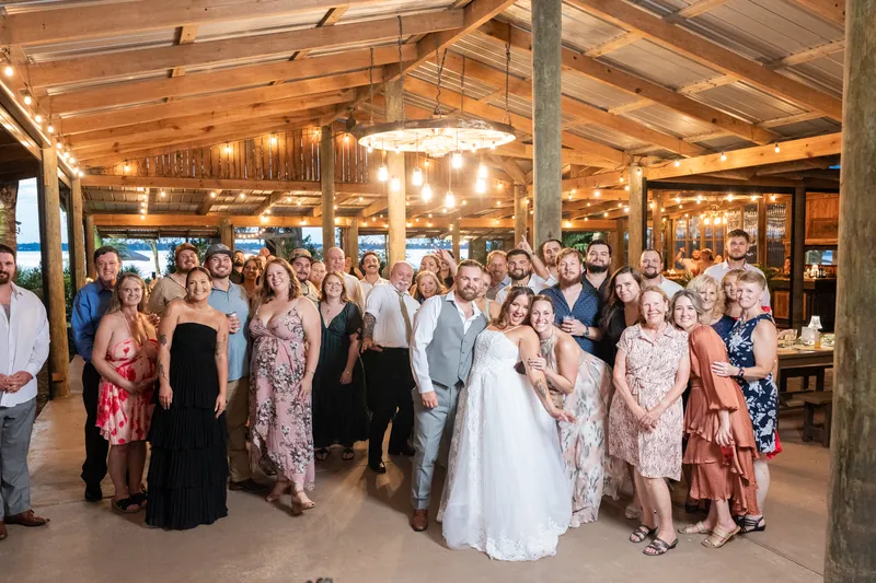 Guests fill a wooden pavilion lit by strung lights, watching the couple celebrate surrounded by family and friends in formal attire.