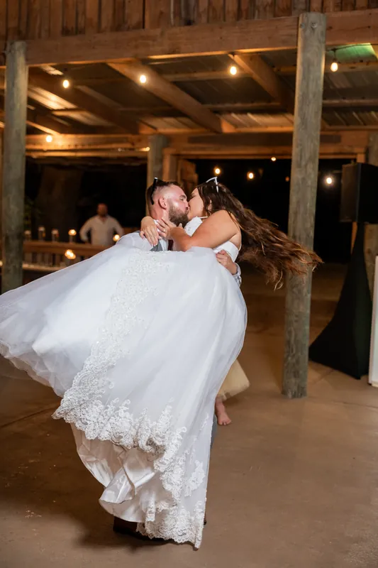 One partner lifts the other in a white gown during their first dance beneath exposed wooden beams and warm overhead lighting.
