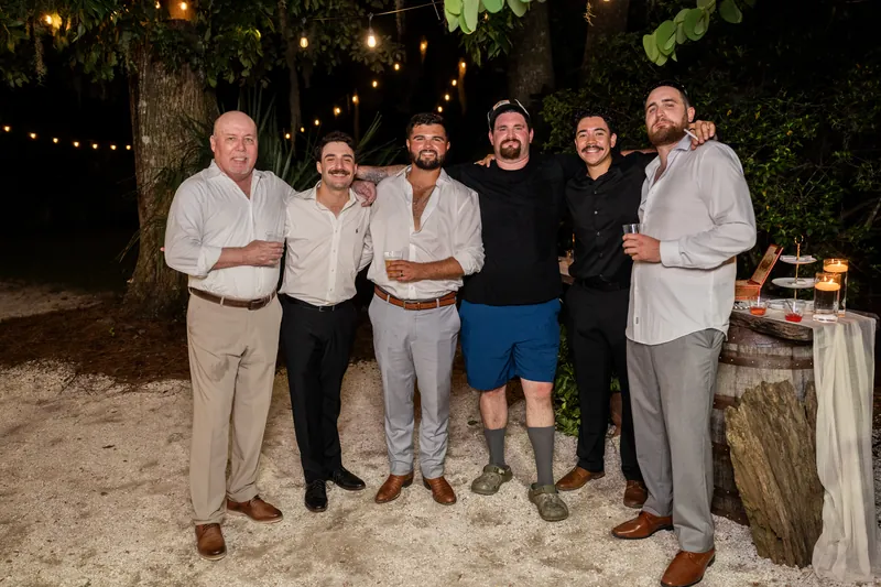 Six men stand shoulder-to-shoulder on sandy ground under market lights, smiling at the camera in a casual groomsmen portrait.