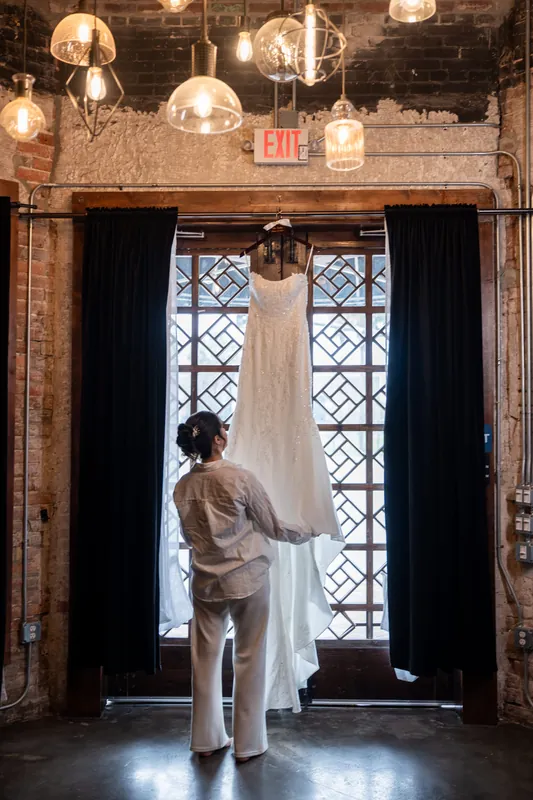 A person in a flowing white gown stands at the entrance of Brick and Beam's industrial brick venue, backlit by tall windows.