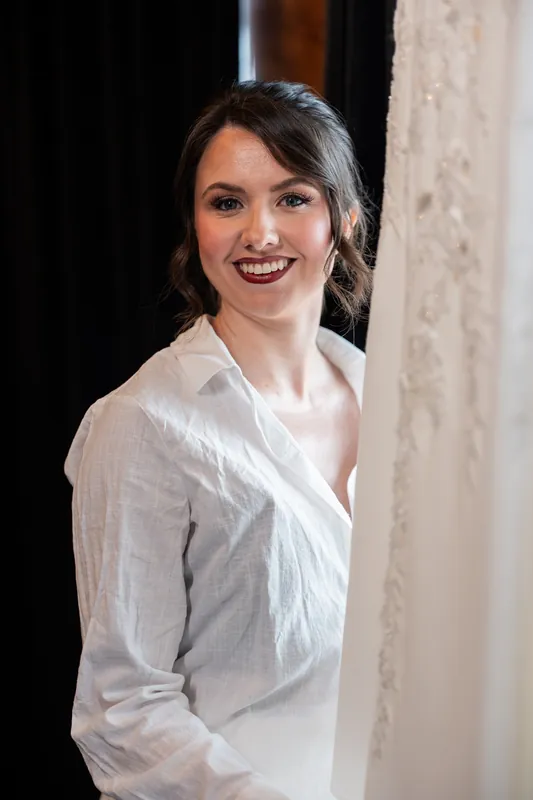 A person with dark hair smiles at the camera wearing a white button-up shirt, standing indoors.