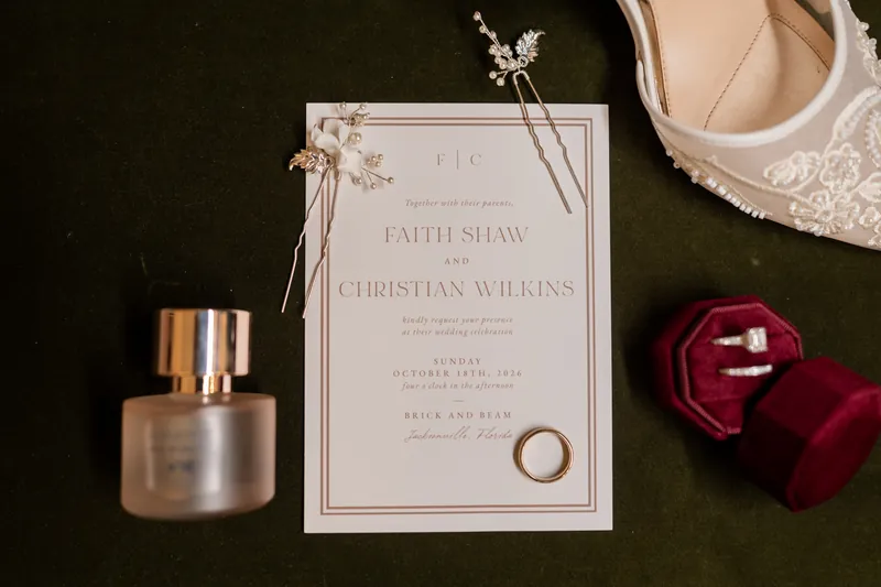 Wedding invitation for Faith Shaw and Christian Wilkins displayed with a perfume bottle, burgundy velvet ring box, and gold band on dark fabric.