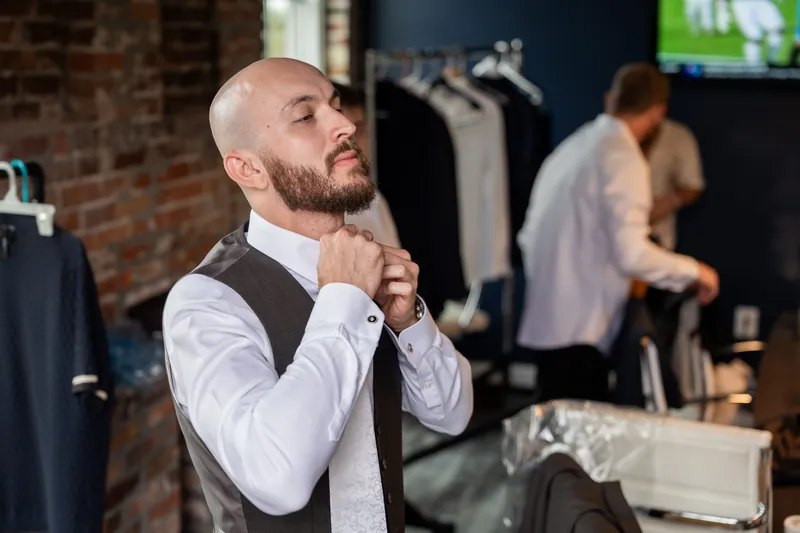 A person in a white dress shirt and vest adjusts a tie while preparing at Brick and Beam, with others visible in the background.