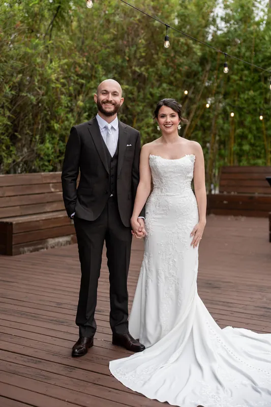 Faith and Christian stand hand-in-hand on a wooden deck, she in a fitted white gown and he in a black suit, with flowering trees behind them.