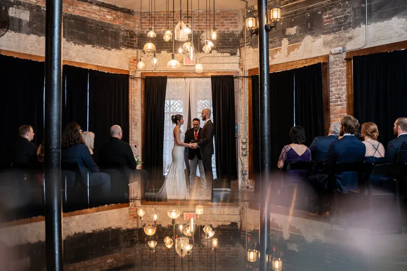 Two people exchange vows beneath hanging pendant lights at Brick and Beam, Jacksonville, with guests seated on either side.