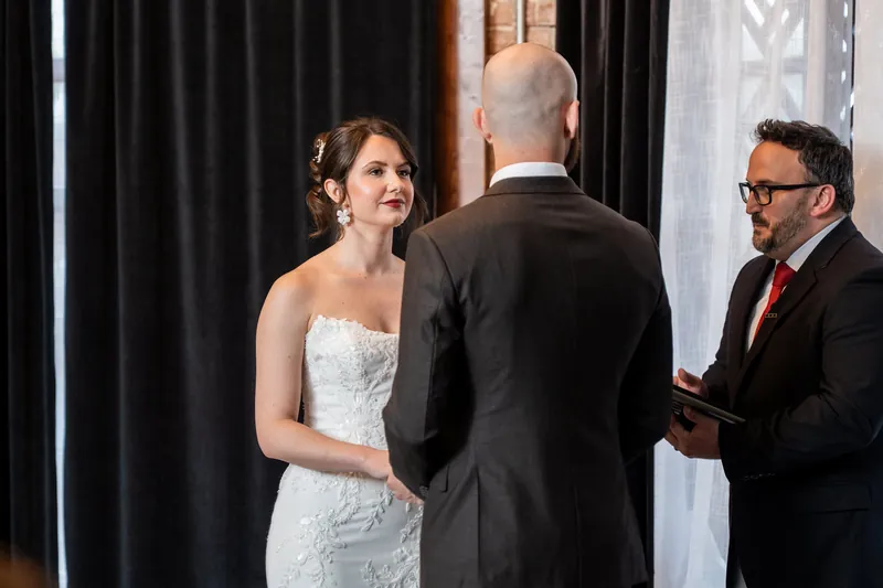 A person in a white gown smiles while speaking with two men in dark suits during the ceremony.