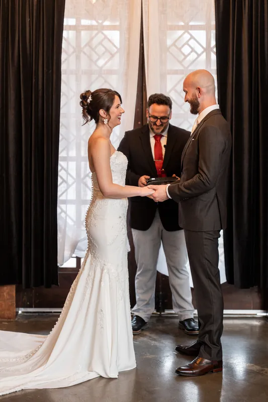 The couple holds hands during their vows as an officiant in a red tie stands between them, with draped windows behind.