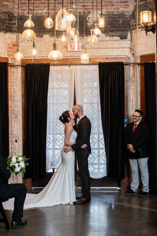 Faith and Christian kiss beneath golden hanging lights at the altar while an officiant watches.