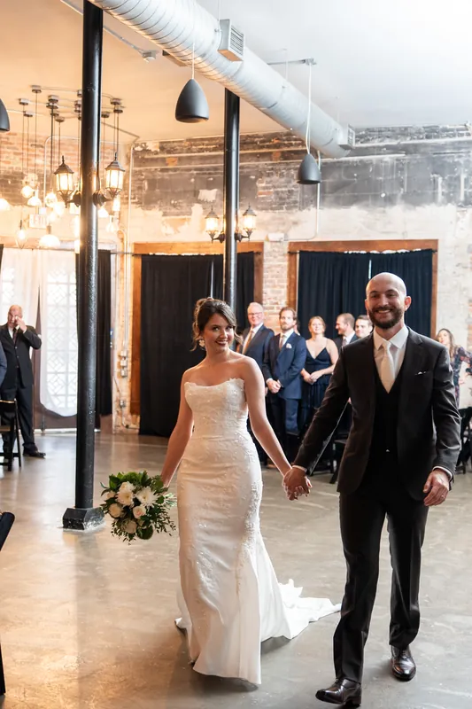 Faith and Christian walk hand-in-hand down the aisle through the industrial brick venue, guests applauding nearby.