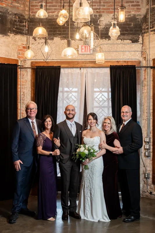 Faith and Christian stand with four family members for a formal portrait in front of the brick wall and illuminated pendant fixtures.