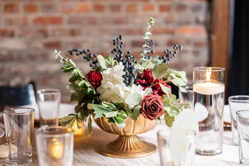 Gold pedestal bowl with cream roses, burgundy blooms, and berries surrounded by water glasses on a dining table.